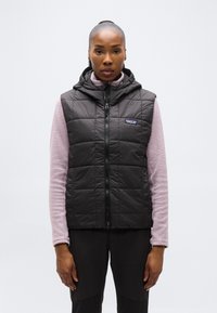 Γυναίκα που φοράει μαύρο, καπιτονέ αμάνικο γιλέκο με κουκούλα πάνω από μωβ ανοιχτόχρωμο, μακρυμάνικο τοπ με υφή, στέκεται μπροστά σε απλό φόντο.