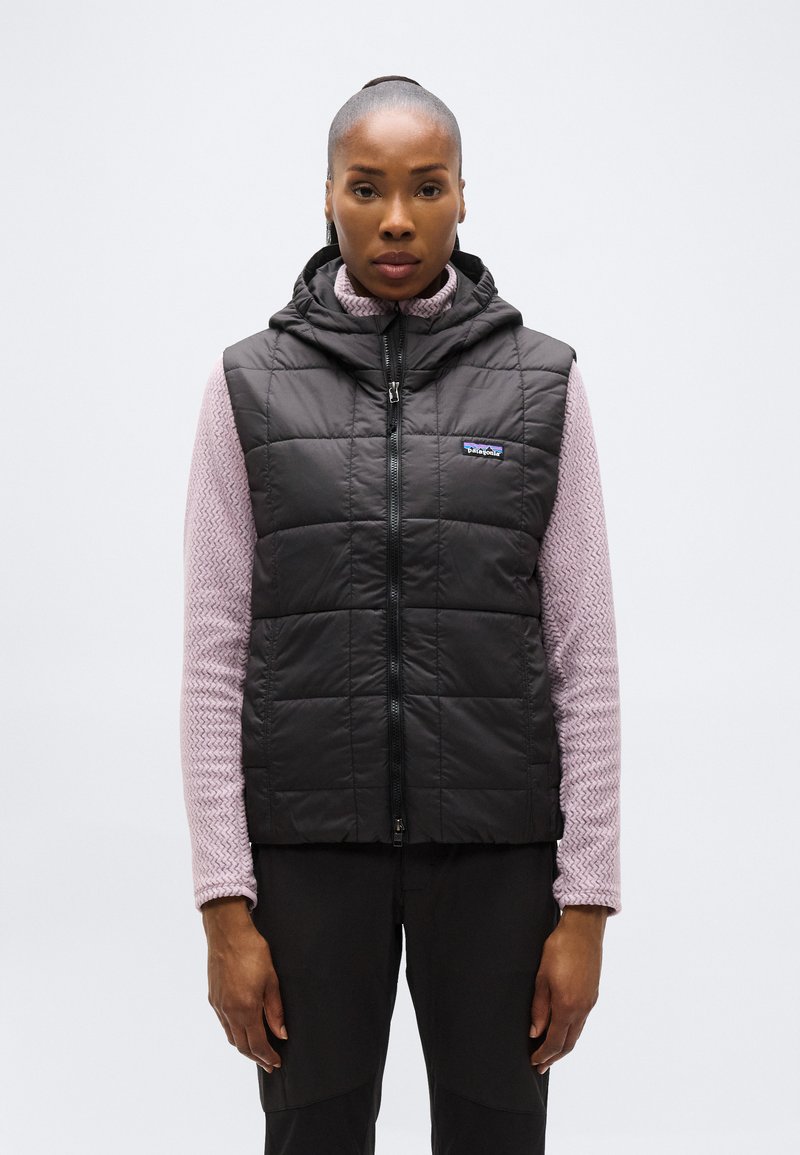 Γυναίκα που φοράει μαύρο, καπιτονέ αμάνικο γιλέκο με κουκούλα πάνω από μωβ ανοιχτόχρωμο, μακρυμάνικο τοπ με υφή, στέκεται μπροστά σε απλό φόντο.