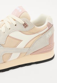 Lys beige sneakers med mesh-overdel og suede-accents i blød pink, hvid logo detalje og en polstret sål med sort mønster.