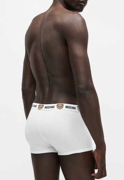 Homme à la peau foncée montré de dos portant un boxer blanc Moschino avec un logo ours sur la ceinture.