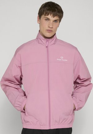 Giovane uomo con capelli castani corti che indossa una giacca rosa chiaro con zip, tasche e logo "Sergio Tacchini" sul petto.