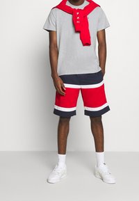 Pánský casual outfit zahrnující šedou košili s krátkým rukávem, červený svetr s kulatým výstřihem uvázaný na krku a šortky s červenými a modrými pruhy. Bílé tenisky.