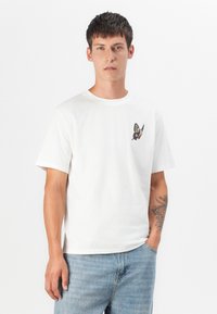 White cotton t-shirt featuring a butterfly embroidery on the left chest, short sleeves, and a round neck; paired with light blue jeans.