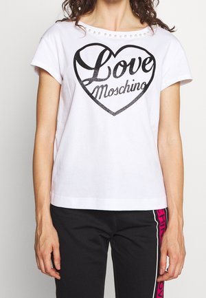 Frau trägt ein weißes T-Shirt mit perlenbesetztem Ausschnitt und schwarzem "Love Moschino" Herz-Print, kombiniert mit schwarzen Hosen mit pinkem Seitentext.