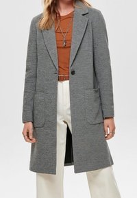 Donna con un lungo cappotto grigio sopra una camicia color ruggine, pantaloni bianchi e una collana con un ciondolo a forma di croce.