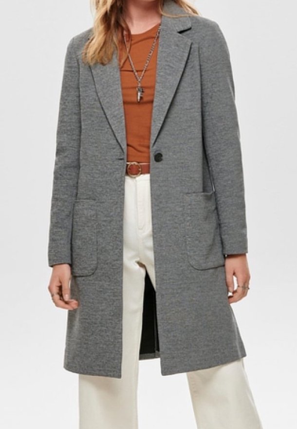 Donna con un lungo cappotto grigio sopra una camicia color ruggine, pantaloni bianchi e una collana con un ciondolo a forma di croce.