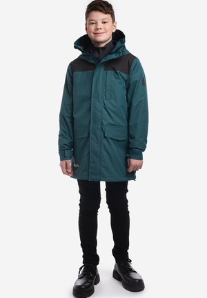 Chłopiec stojący w turkusowo-czarnej parce z kapturem, czarnych spodniach i czarnych butach na białym tle.