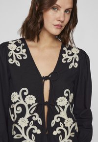 Blusa preta com um decote em V profundo, apresentando um intrincado bordado floral branco e um fecho de laço na frente, mangas longas e volumosas.