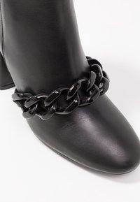 Botín negro de cuero suave con punta redondeada y un llamativo detalle de cadena negra gruesa alrededor del tobillo.