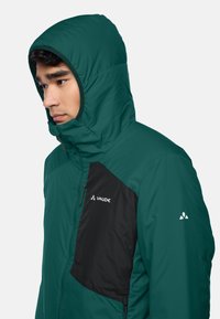 Grüne isolierte Jacke mit schwarzem Brustpanel, vorderem Reißverschluss und verstellbare Kapuze. Mit Logo auf der Brust und dem Ärmel. Glatte Textur.