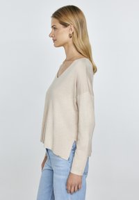 Beige, V-nyakú pulóver hosszú ujjakkal, puha textúrával és oldalsó hasítékokkal. Világoskék farmernadrággal párosítva, bemutatva egy laza sziluettet.