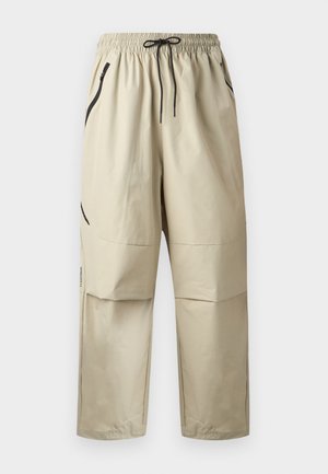 Pantalon léger beige avec taille élastique, cordon de serrage noir et poches latérales zippées, présenté de face sur un fond uni.