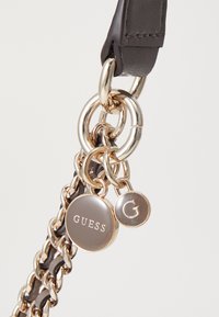 Black leather strap featuring gold-tone chain accents and logo charms displaying "GUESS" and "G." Smooth texture with polished hardware.