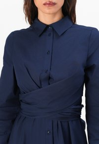 Mörkblå skjortklänning med långa ärmar, krage, knäppning fram och en detalj i knytstil vid midjan. Slät tyg med draperad accent.