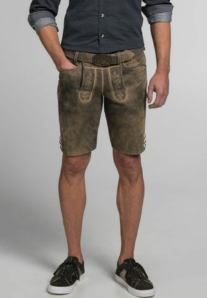 Lederhosen in einem verwitterten Braunton mit gelben Nähten; ausgestattet mit Taschen und dekorativen Knöpfen an der Seite. Kombiniert mit schwarzen Sneakers.