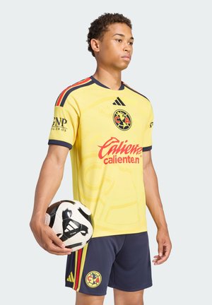 Junger männlicher Fußballspieler in gelber und marineblauer Uniform, der einen schwarz-weißen Adidas-Fußball an seiner Seite hält.