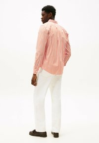 Camicia a maniche lunghe a righe rosse e bianche con colletto abbottonato, abbinata a pantaloni bianchi e scarpe scure. Tessuto morbido, design aderente.