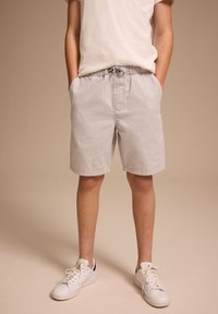 Persona che indossa pantaloncini grigio chiaro con coulisse, scarpe da ginnastica bianche e una maglietta bianca, in piedi con le mani in tasca contro uno sfondo beige.