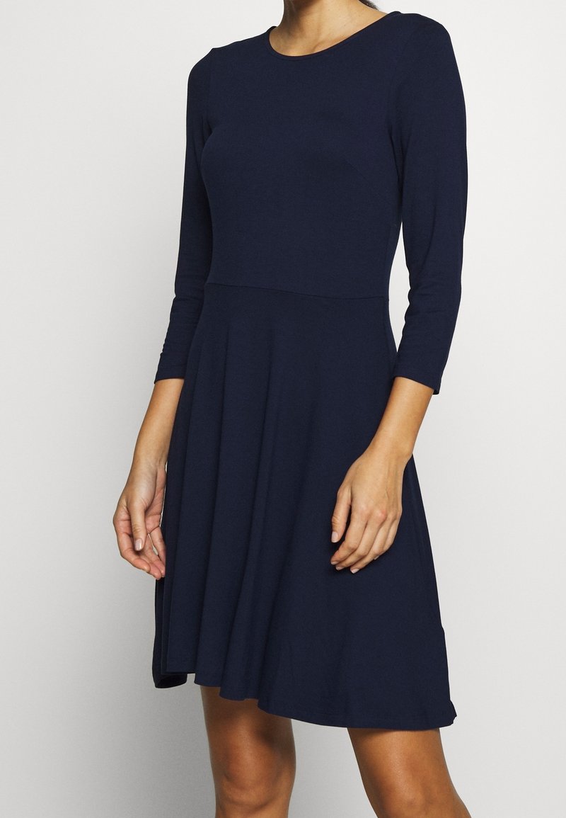 Femme portant une robe bleu marine longueur genou avec des manches trois-quarts et une taille cintrée.