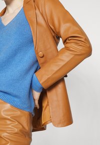 Orange Lederblazer mit einem einzigen Knopf, über einem hellblauen Wollpullover mit strukturierten Oberflächen, kombiniert mit passenden Hosen.