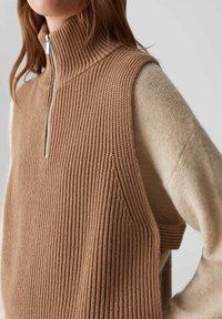 Beige gestrickte Weste mit hohem Rippenkragen und einem Reißverschluss. Unter der Weste wird ein hellbrauner, langärmeliger, strukturierter Pullover hervorgehoben.