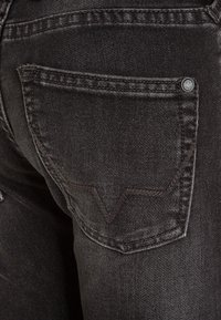 Jeans de mezclilla negra con un bolsillo trasero que presenta un detalle de costura curva y un botón metálico. La tela tiene una textura ligeramente desteñida.