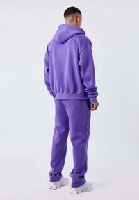 Hombre de pie de espaldas, vistiendo un conjunto a juego de sudadera y pantalones deportivos morados, con zapatillas blancas, sobre un fondo liso.