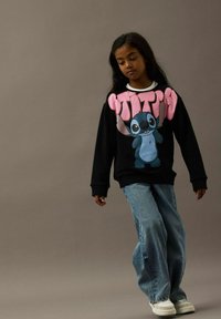 Ragazza con lunghi capelli scuri che indossa un felpa nera con un personaggio dei cartoni e testo rosa, abbinata a jeans blu larghi e sneakers bianche.