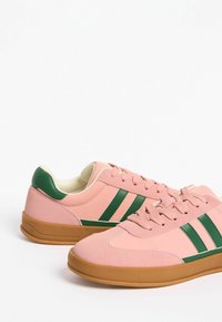 Rozā sporta apavi ar zaļām svītrām, gumijas zolēm un apaļu formu. Teksturēts augšdaļas materiāls ar šņoru detaļām un mīkstinošu iekšpusi.