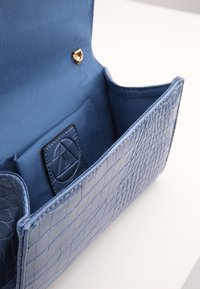 Modrá kožená kabelka s krokodýlí texturou z vnější strany, zlatým drukem a hladkým saténovým vnitřkem s logem.