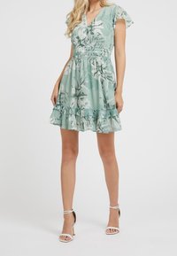 Robe à manches courtes, couleur vert menthe, avec motif floral, taille smockée et ourlet volanté. Associée à des sandales à talons hauts blanches avec des lanières.