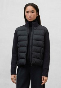 Chaleco acolchado negro con collar alto, con cierre de cremallera y acolchado horizontal. Lleva puesto sobre un suéter oscuro. Textura suave y diseño ajustado.