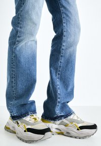 Teknős sneakers, szürke és fekete hálós felsőrésszel, sárga akcentussal, texturált talppal és széles szilhuettel, világoskék farmernadrággal párosítva.