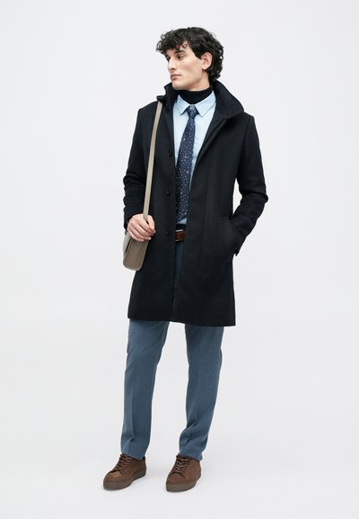Cappotto nero in misto lana con cappuccio, indossato sopra una camicia azzurro chiaro e una cravatta a fantasia. Pantaloni grigi e scarpe marroni con lacci completano il look.