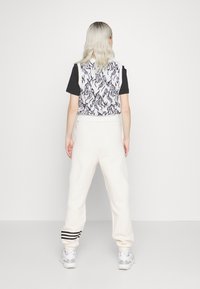 Pletená bílá vesta s černým abstraktním vzorem, nošená přes černé tričko. Bílé tepláky s černými pruhy na manžetách, šedé tenisky.