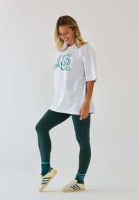 Biały, oversize'owy t-shirt z zielonym graficznym napisem, w połączeniu z ciemnozielonymi legginsami i beżowymi sneakersami z ciemnymi paskami. Koszulka ozdobiona gwiazdami.