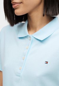Femme portant un polo bleu clair à quatre boutons blancs et un petit logo rectangulaire rouge, blanc et bleu sur la poitrine.