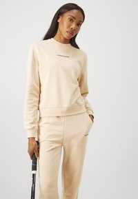 Beige twee-delige outfit van zachte stof, bestaande uit een sweatshirt met een ronde hals en bijpassende broek met zijzakken. Groene logodetail.