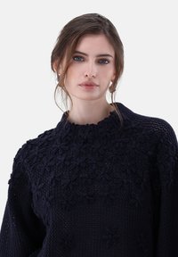 Junge Frau mit heller Haut und blauen Augen, die einen dunkelblauen Strickpullover mit floralen Mustern trägt, ihre Haare locker gebunden mit Strähnen, die ihr Gesicht umrahmen.