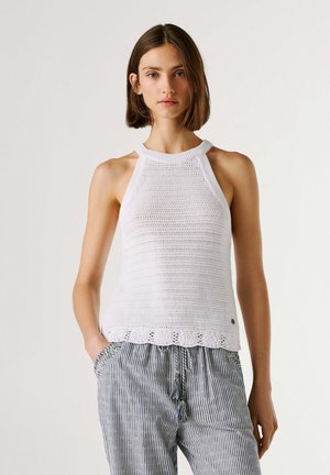 Femme portant un haut à bretelles en maille blanche sans manches et un pantalon ample rayé, les mains dans les poches, debout devant un fond clair uni.