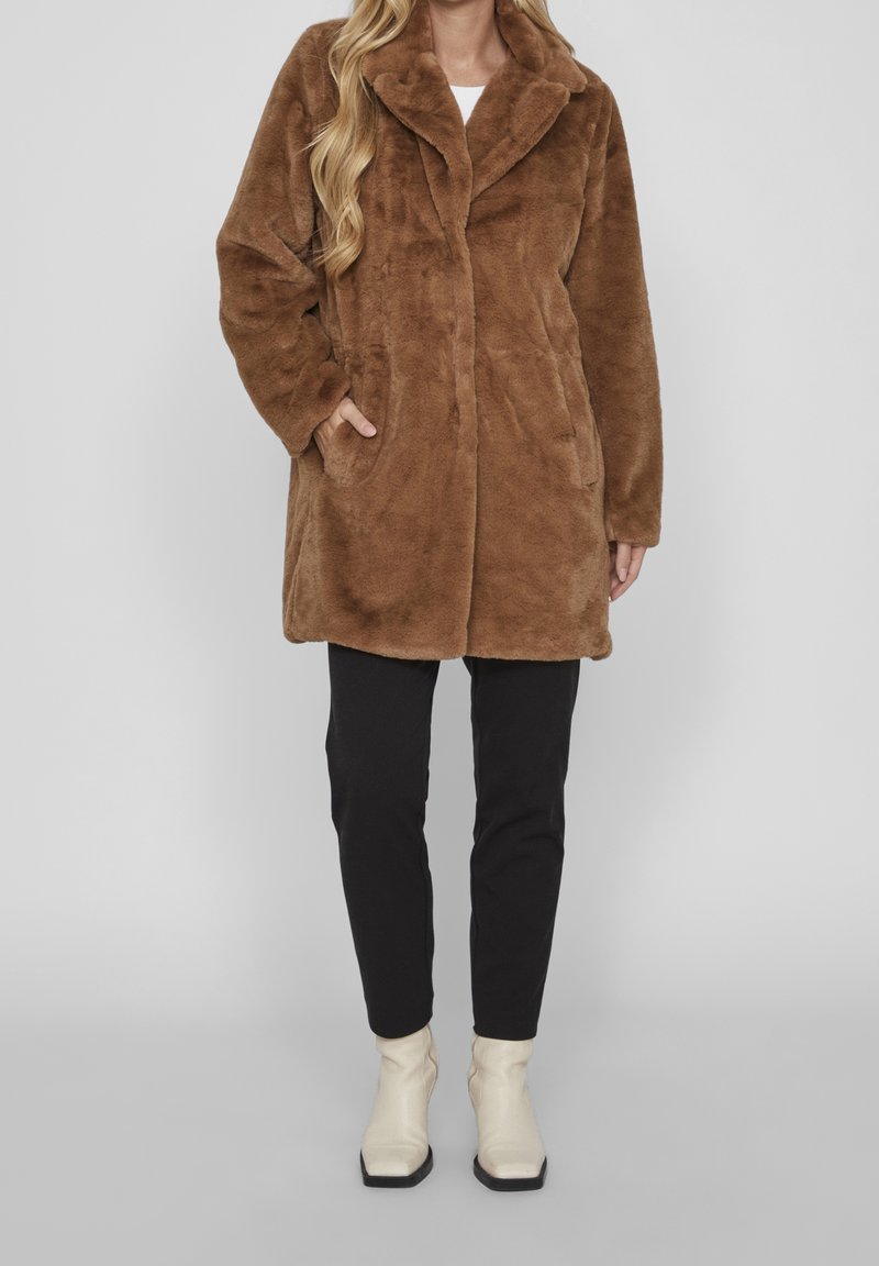 Femme portant un manteau mi-long en fausse fourrure marron, un pantalon noir et des bottines blanches, debout devant un fond gris uni.