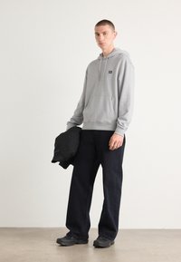 Grijze katoenen hoodie met een voorzak en een capuchon met trekkoord, gecombineerd met donkerblauwe wijde broek en zwarte schoenen, gedragen met een zwart jack.