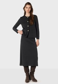 Veste en tricot noire avec des boutons dorés et deux poches, associée à une longue jupe noire. Portée avec des bottes marron foncé jusqu'aux genoux.