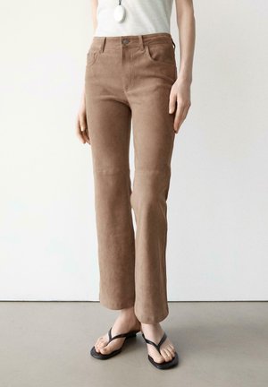 Persona che indossa pantaloni marroni in camoscio a vita alta e gamba dritta, con una canotta bianca senza maniche e sandali infradito neri, in piedi su un pavimento chiaro.