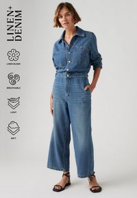 Combinaison en denim bleu clair avec un mélange de lin. Elle dispose d'un devant boutonné, d'une taille élastique, de deux poches poitrine et d'un design à jambes larges.