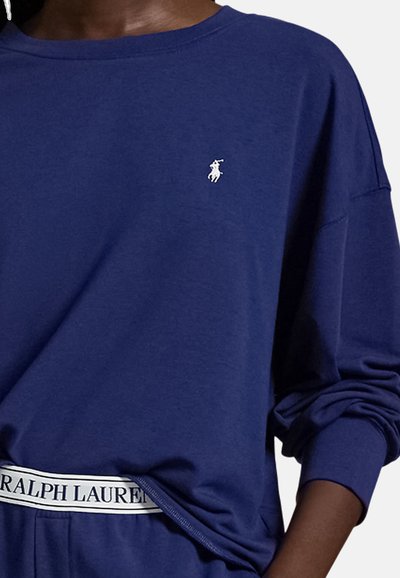 Άνδρας που φοράει μπλε σκούρο πουκάμισο με μακριά μανίκια και μικρό λευκό λογότυπο παίκτη πόλο, και παντελόνι που ταιριάζει με εμφανές λάστιχο μέσης Ralph Lauren.