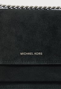 Bolso de mano de ante negro con correa de cadena plateada y logo en relieve "MICHAEL KORS" en la parte frontal. Acabado texturizado y suave con un diseño elegante.