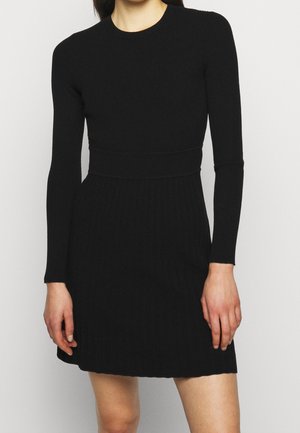 Kobieta ubrana w czarną, długorękawową sukienkę z prążkowanego dzianiny, dopasowaną w talii i z krótkim dołem powyżej kolan.