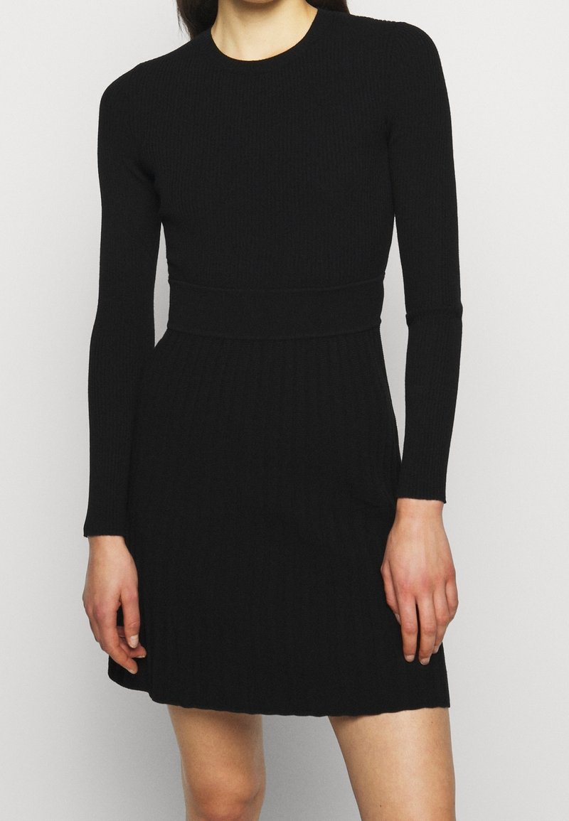 Femme portant une robe noire en maille côtelée à manches longues, avec une taille cintrée et un ourlet court au-dessus des genoux.