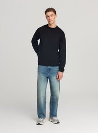 Maglione nero in maglia abbinato a jeans blu slavati. Il modello indossa sneakers grigie, caratterizzate da una vestibilità comoda e dettagli minimalisti in tutto il capo.